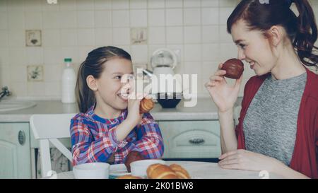 Buona madre e simpatica figlia a colazione mangiando muffin e parlando a casa in cucina moderna. Concetto di famiglia, cibo, casa e persone Foto Stock