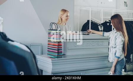 Allegra cliente femminile che si trova alla scrivania del cassiere, che chiacchiera con il venditore e paga gli acquisti con lo smartphone. Foto Stock