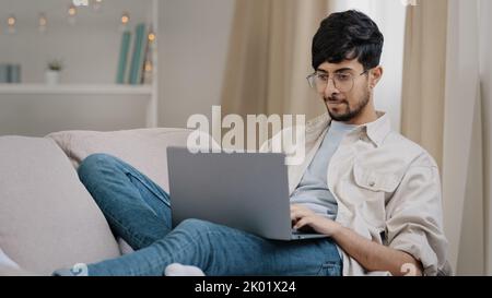 Giovane ragazzo arabo freelance uomo d'affari maschio barbuto studente in occhiali si trova sul divano a casa con il computer portatile che lavora a studiare la lettura online pensiero Foto Stock