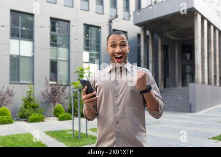 Felice e scioccato dalle notizie di mattina, l'uomo d'affari arfo-americano sorridente e guardando la macchina fotografica dall'esterno dell'edificio dell'ufficio, l'uomo che cammina giù per strada usando il telefono mobile. Foto Stock