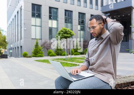 Uomo serio e penoso seduto sul banco insoddisfatto del risultato del lavoro, utilizzando il laptop per il lavoro remoto, uomo d'affari afro-americano fuori dall'ufficio edificio seduto sul banco. Foto Stock