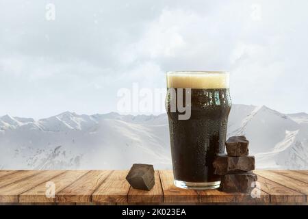 Cioccolato fondente e birra. Grande bicchiere di birra scura e schiumosa che si erge su un tavolo di legno sullo sfondo delle montagne innevate. Foto Stock