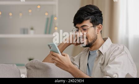 Primo piano arabo ragazzo ispanico giovane uomo che indossa occhiali bearded maschio utente blogger seduto a casa con il telefono cellulare chattare online navigazione web digitando Foto Stock