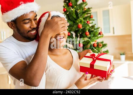 afro americano uomo che dà scatola regalo alla sua ragazza in casa vicino albero di natale Foto Stock