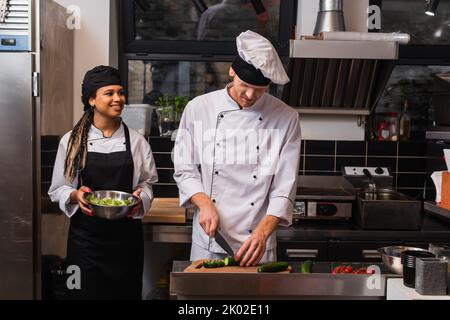 donna afroamericana in grembiule tenere ciotola con lattuga e guardando chef preparare pasto in cucina, immagine stock Foto Stock