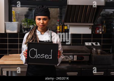 giovane cuoco africano americano che tiene la lavagna con la scritta chiusa in cucina, immagine di scorta Foto Stock