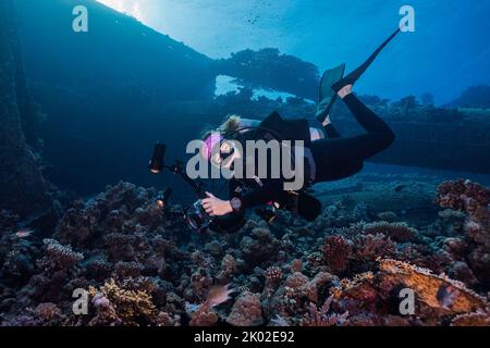 Un fotografo subacqueo che guarda la macchina fotografica scattando foto sulla barriera corallina con un relitto sullo sfondo Foto Stock