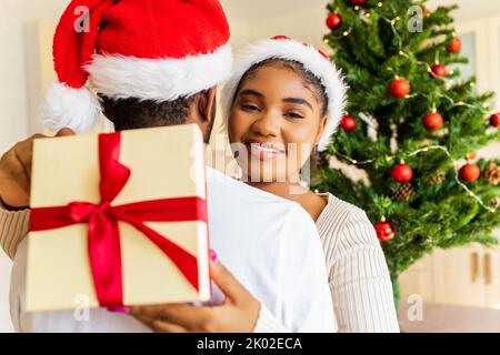 afro american coppia che dà scatola del regalo l'un l'altro in casa vicino all'albero di natale Foto Stock
