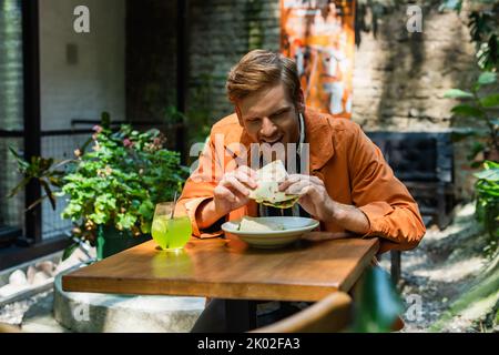 allegro uomo rosso mangiare quesadilla vicino al vetro con cocktail in terrazza all'aperto, immagine stock Foto Stock