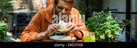 allegro uomo rosso mangiare quesadilla vicino al vetro con cocktail in terrazza all'aperto, banner, immagine stock Foto Stock