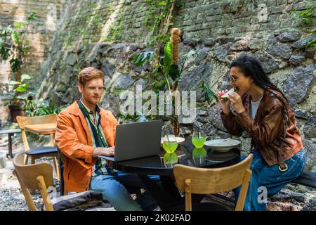 afroamericana donna mangiare quesadilla vicino freelancer amico utilizzando il portatile in terrazza all'aperto, immagine stock Foto Stock