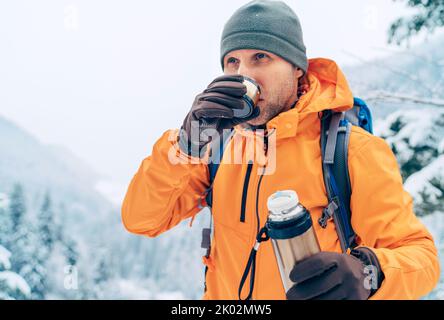 Uomo che beve una bevanda calda da thermos fiasca vestito giacca softshell arancione brillante mentre lui trekking montagna percorso invernale. Persone attive nel natu Foto Stock