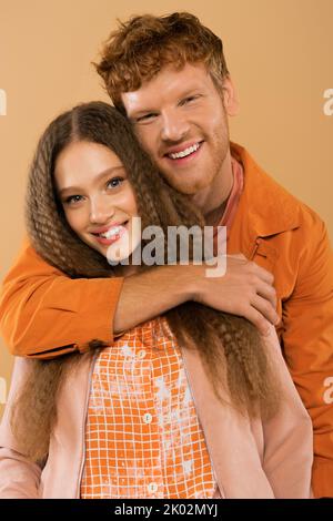 allegro giovane uomo rosso che abbraccia felice ragazza con capelli ondulati isolato su beige, immagine stock Foto Stock