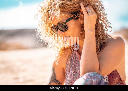 Donna bella di mezza età con capelli ricci e occhiali da sole in esterno ritratto laterale. Moderna e simpatica donna con tatuaggio rilassarsi in spiaggia sotto il sole estivo. Vita sana delle persone bionde Foto Stock