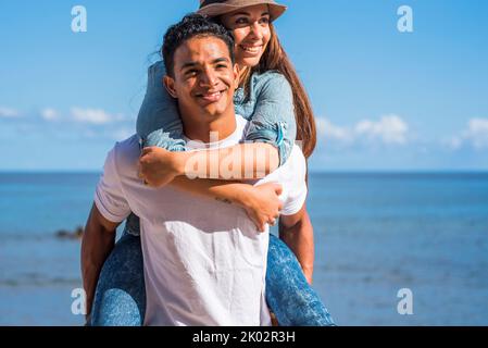Felice giovane coppia nera e caucasica godendosi e divertendosi insieme in rapporto all'aperto. Incontri e attività di svago persone. L'uomo porta la donna sulla schiena. Blu oceano e cielo sfondo turistico Foto Stock