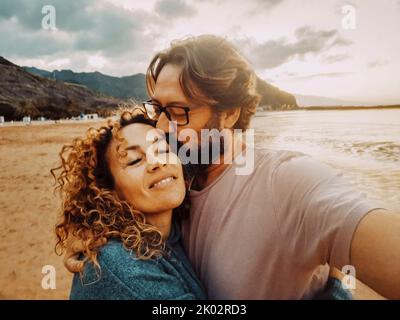 Felice coppia adulta innamorata godersi vacanze estive insieme a scattare selfie foto con il telefono cellulare. Uomo che bacia donna sorridente. Uomo e donna che abbraccia la spiaggia Foto Stock
