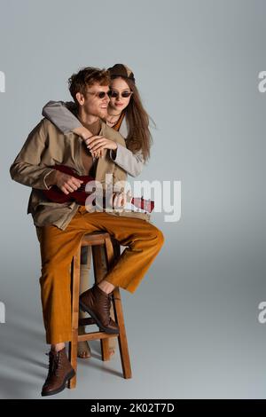 felice giovane uomo giocando ukulele vicino ragazza mentre seduto su sedia alta sul grigio, immagine stock Foto Stock