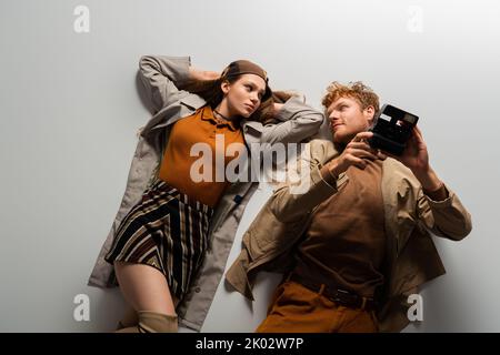 vista dall'alto di un giovane uomo rosso in vestito autunnale con fotocamera vintage e sdraiato insieme alla ragazza su immagine grigia, stock Foto Stock