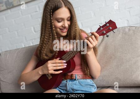 gioiosa ragazza adolescente in abiti casual giocando ukulele mentre si siede sul divano in soggiorno, immagine stock Foto Stock