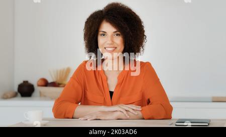 Webcam vista sorridente afroamericana donna con capelli ricci seduta a casa cucina blogger femmina influenzatore vlogger ondulazione mano saluto Foto Stock