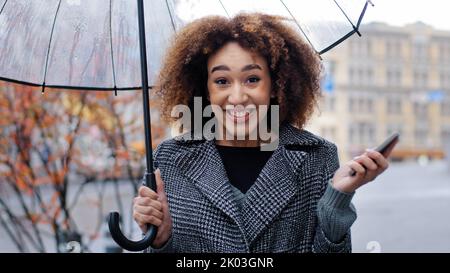 Ragazza afroamericana donna con ombrello trasparente stand in città sotto la pioggia guardare divertente video sul telefono riceve umoroso messaggio ride forte Foto Stock