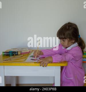 adorabile bambina in grembiule rosa, seduta su una scrivania mentre colorava un notebook con colori a matita. Ritorno a scuola e attività svolte da kin Foto Stock