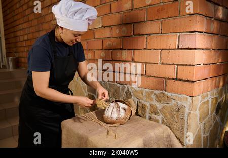 Donna panettiere posti spikelets di grano accanto a pane fresco cotto a grani interi su tavola coperta di tovaglia di lino Foto Stock