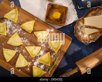 Ravioli grezzi triangolari su un tagliere infarinato. Cibo sfondo. Ingredienti, utensili da cucina. Molti oggetti. Vista ad angolo basso. Ricette di italiano Foto Stock