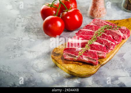 Bistecche di carne cruda su un tagliere e pomodori freschi maturi su sfondo grigio chiaro. Ricette per piatti a base di carne di ristorante e cucina casalinga. Banchetto Foto Stock