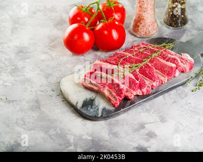 Pezzi di carne cruda selezionata su un tagliere con un rametto di rosmarino, pomodori maturi e spezie su sfondo chiaro. Ricette per cucinare piatti wi Foto Stock