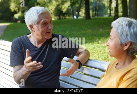 Coppia di mezza età sposi multi-etnici siedono riposo sulla panchina godere parlare nel parco estivo pubblico avere conversazione amichevole condividere pensieri o notizie durante Foto Stock