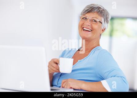 Donna che si diverte a bere un caffè mentre usa un computer portatile. Donna matura sorridente che si diverte a gustare il caffè mentre usa il notebook a casa. Foto Stock