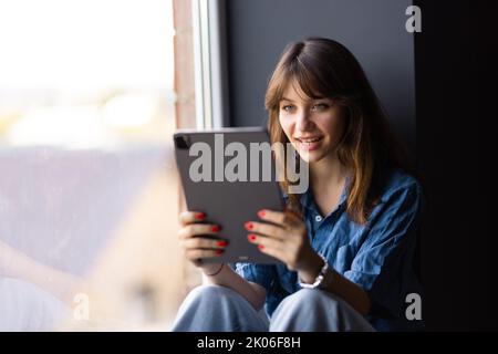 Triste scioccato giovane donna adolescente utilizzando tablet vedere qualcosa di sgradevole in internet seduta sul windowsill. Foto Stock