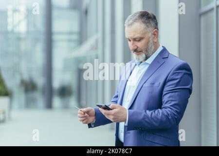 Un uomo d'affari di sesso maschile serio e focalizzato è in piedi vicino a un centro di uffici, tenendo un telefono cellulare e una carta di credito in mano, inserendo il numero di carta nel conto sul telefono. Foto Stock