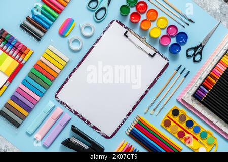 Forniture scolastiche colorate disposte su sfondo blu con carta comune bianca al centro Foto Stock