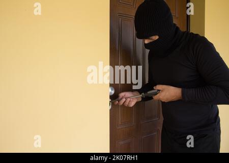 Immagine di un ladro vestito tutto nero con una balaclava che cerca di forzare la serratura di una porta di casa con un grosso cacciavite. Aumento costante in casa Foto Stock