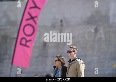 Giovane coppia in piedi sulla spiaggia di guardare la concorrenza di surf con la bandiera di pubblicità di Roxy al concorso di surf Foto Stock