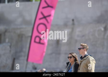 Giovane coppia in piedi sulla spiaggia di guardare la concorrenza di surf con la bandiera di pubblicità di Roxy al concorso di surf Foto Stock