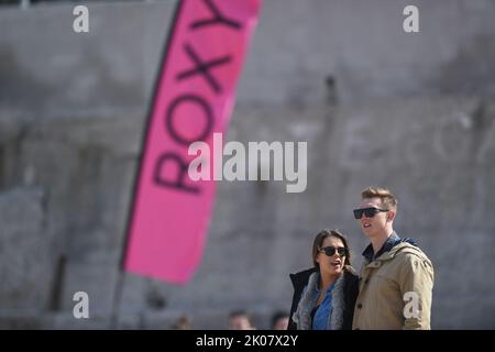 Giovane coppia in piedi sulla spiaggia di guardare la concorrenza di surf con la bandiera di pubblicità di Roxy al concorso di surf Foto Stock