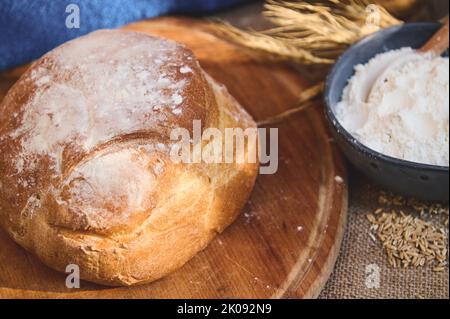 Una pagnotta di pane a pasta madre su una tavola di legno, contro una ciotola di ceramica blu sfocata con farina bianca e spikelets di grano Foto Stock