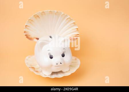 Piggybank bianco in una conchiglia di mare di ostriche su fondo arancione molto morbido. Concetto esotico di risparmio di vacanza, fuoco morbido primo piano Foto Stock