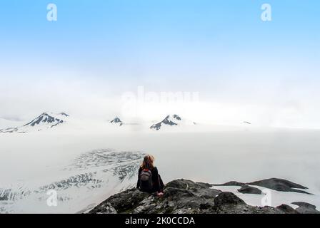 Escursionista femminile seduto in cima a una cima di montagna che si affaccia sul campo di ghiaccio Harding in Alaska, ammirando gli incredibili panorami e panorami surreali Foto Stock