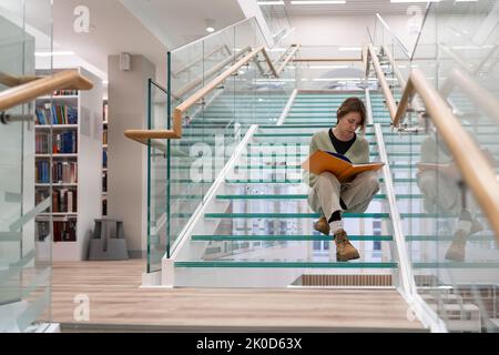 Curioso lettore femminile si siede su scale di vetro moderno passo lettura libro preferito sulla storia del mondo Foto Stock