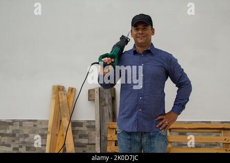 sorridente handyman con un hacksaw sulla spalla mentre taglia e smonta pallet e piattaforme di legno. Riutilizzo di legno per fare-it-yourself lavori Foto Stock