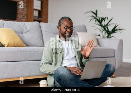 Felice manager afro-americano maturo maschio in occhiali e casual mano ondulata, guarda il pc, sedersi sul pavimento Foto Stock