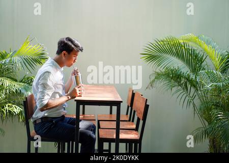 Un uomo in camicia bianca azzurro era seduto ad un lungo tavolo marrone, bevendo acqua di paglia, ci sono palme gialle stand accanto e di fronte a lui. Foto Stock