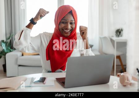 Ottime notizie. Entusiasta musulmana afro-americana festeggia il successo, guardando lo schermo del notebook, alzando pugni ed esclamando Foto Stock