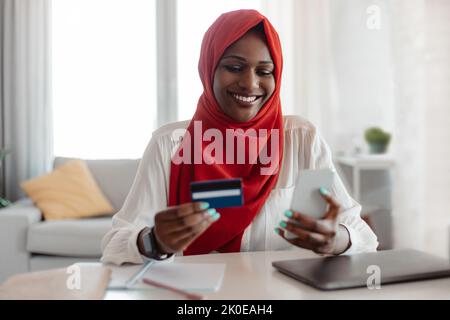 Felice afroamericana donna in hijab utilizzando smartphone e carta di credito, shopping sul web a casa Foto Stock