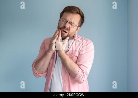 L'uomo con gli occhiali sta sperimentando un forte mal di denti, mantiene la mano sulla guancia e chiude gli occhi nella sofferenza. Concetto medico. Foto Stock