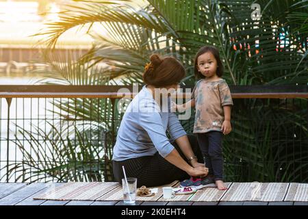 BANGKOK,THAILANDIA-FEBBRAIO 23,2020:nonna non identificata si prende cura di nutrire il suo nipote cibo e acqua mentre si rilassa sulla terrazza lungo il fiume a Hi Foto Stock